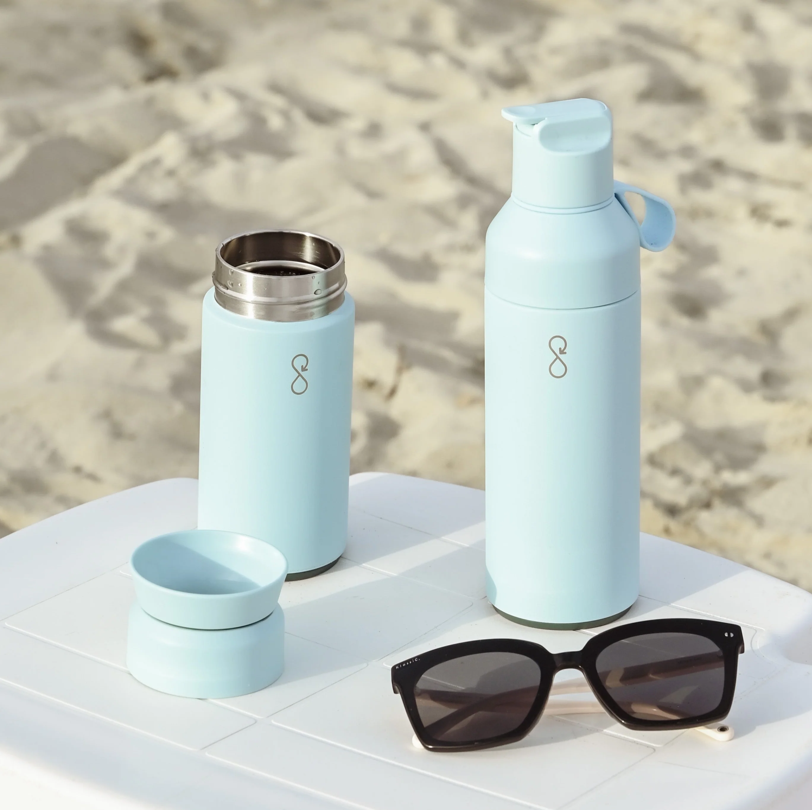 A pair of light blue Ocean Bottles with different lids on a table on the beach